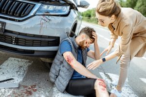 A man sitting on the floor holding his head after a car accident with a woman helping him.