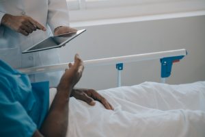 A patient is lying down in bed and has suffered medical negligence. A health care professional stands at their bedside pointing to a tablet